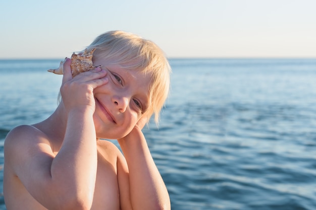 Chico rubio lindo puso concha a la oreja en el mar. Vacaciones en el mar con niños.