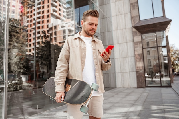 Chico rubio milenario enfocado durante la publicación de un teléfono inteligente que le gusta la transmisión de pie en el fondo de la calle urbana Estudiantes que se comunican a través de un dispositivo de teléfono celular, un patinador inconformista con longboard