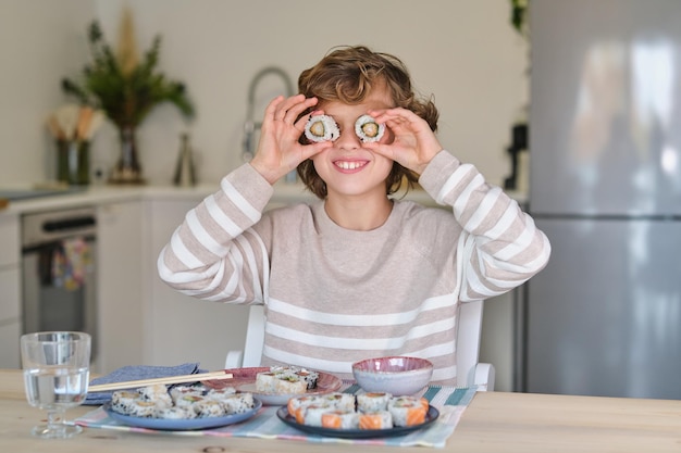 Chico rubio positivo divirtiéndose con rollos mientras está sentado en la mesa con un plato de sushi sabroso en la cocina ligera
