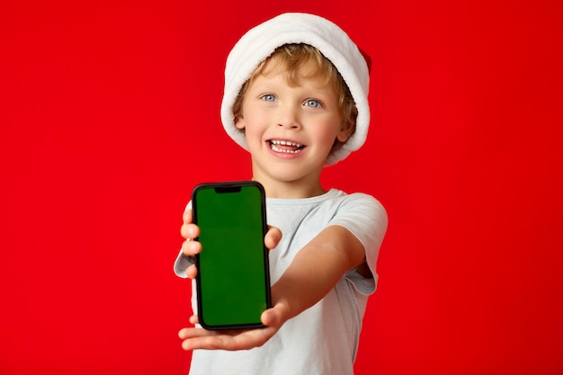 Chico rubio sonriente en un divertido fondo de gorra de Navidad muestra la pantalla del teléfono en blanco