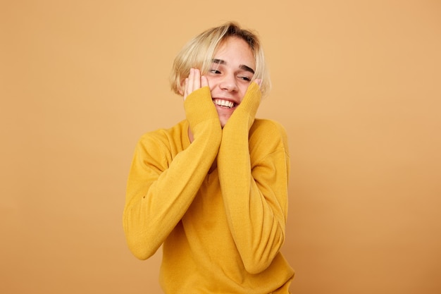 Chico rubio vestido con suéter amarillo mantiene sus manos en su rostro sobre el fondo beige en el estudio.