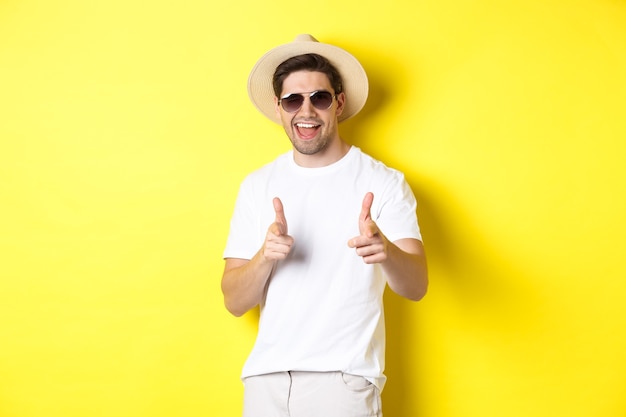 Foto chico seguro y descarado de vacaciones coqueteando contigo, señalando con el dedo al frente y guiñando un ojo, con sombrero de verano con gafas de sol, pared amarilla