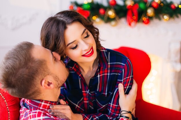 El chico sentado en el sofá y abraza a su novia y quiere besarla y ella cerró los ojos en el espacio decorado con chimenea.