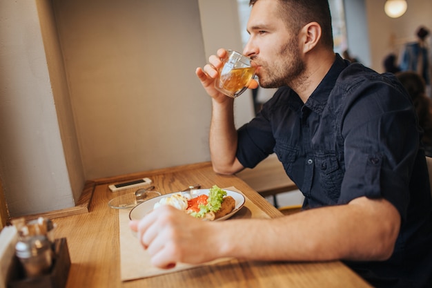 Chico serio está sentado a la mesa y tomando té. Él está mirando hacia adelante. También hay un plato con gofres y verduras.