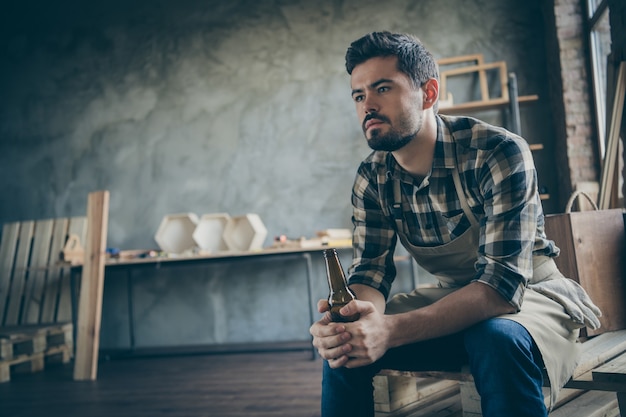 chico serio guapo sosteniendo una botella de cerveza tiene que trabajar en el extranjero para ganar dinero señorita familia final del día solitario industria de madera tienda de carpintería en el interior