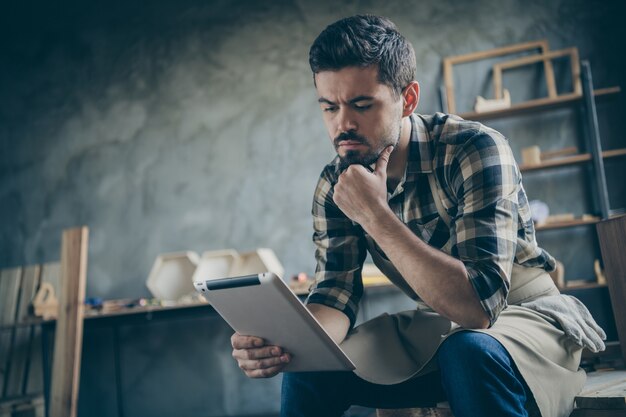 chico serio guapo sosteniendo e-reader sitio web revisando el correo electrónico pedidos en línea piense cómo mejorar el servicio negocio de madera propietario de la industria tienda de carpintería en el interior