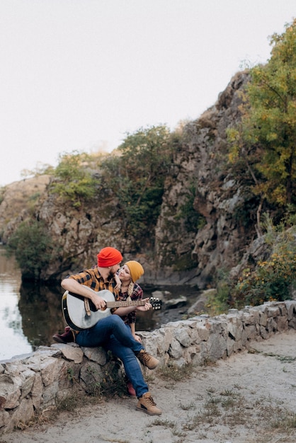 Un chico con un sombrero brillante toca la guitarra con una chica sobre un fondo de rocas de granito y un río