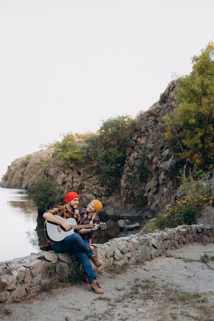 Un chico con un sombrero brillante toca la guitarra con una chica sobre un fondo de rocas de granito y un río