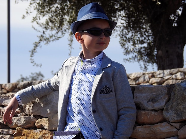 Chico con sombrero y gafas de sol posando junto a un muro de piedra frente a un árbol gigante