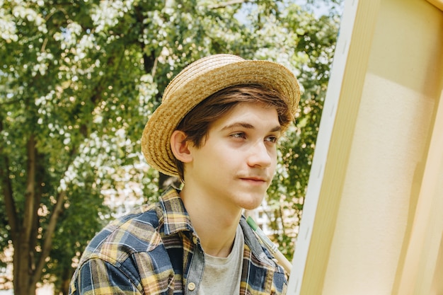 chico con sombrero de paja se sienta en el parque frente a un caballete