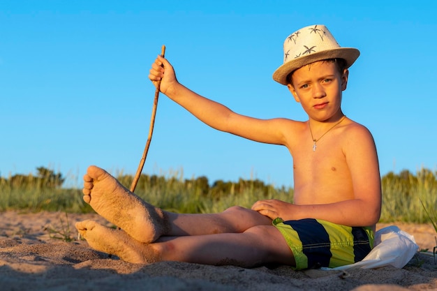 El chico con sombrero para el sol y pantalones cortos de baño se relaja sentado en la playa