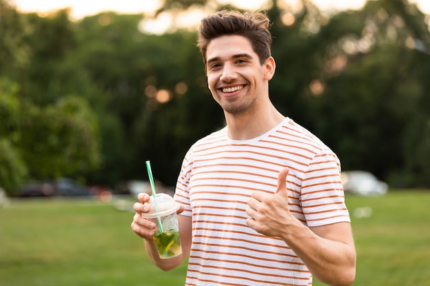 Chico sonriente caminando por el parque y bebiendo bebidas de un vaso de plástico