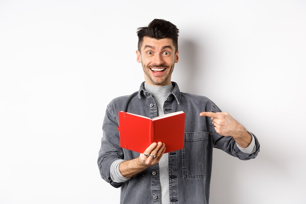 Chico sonriente feliz apuntando al planificador rojo, mostrando un buen diario o libro, de pie sobre fondo blanco.