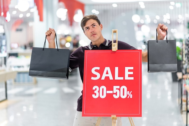 Chico sonriente joven posando con una camiseta burdeos y pantalones cortos beige con bolsas de colores. posando en el fondo de la pancarta con la inscripción venta y descuentos de hasta 30-50 por ciento