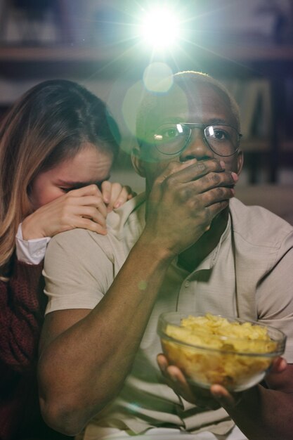 Chico sorprendido de etnia africana cubriendo la boca con la mano mientras sostiene el tazón con papas fritas, su novia molesta poniendo la cabeza en su hombro