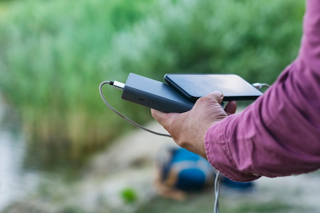 Foto el chico sostiene un cargador portátil con un teléfono inteligente en la mano. hombre sobre un fondo de naturaleza con vegetación y un lago.