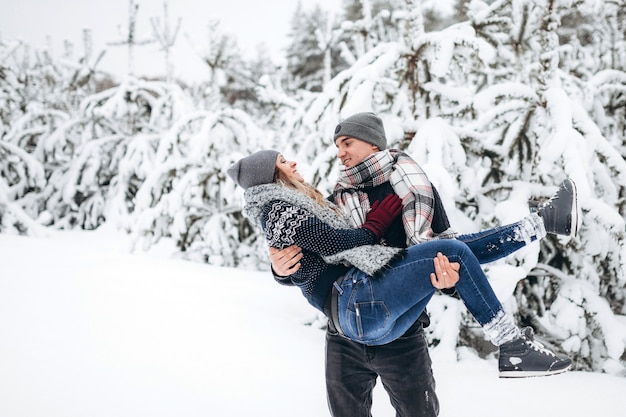 El chico sostiene a la chica en sus brazos en el bosque nevado de invierno