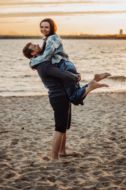 El chico sostiene a la chica en sus brazos en la playa. Cita romántica de verano al atardecer.