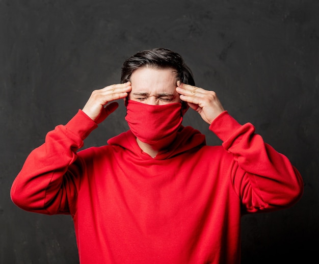 Chico en sudadera roja y mascarilla con dolor de cabeza en la pared oscura