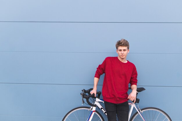 Chico en una sudadera roja de pie con una bicicleta blanca en el fondo de una pared azul