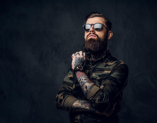 Chico tatuado barbudo en camisa militar y gafas de sol posando con mirada pensativa. Foto de estudio contra una pared oscura