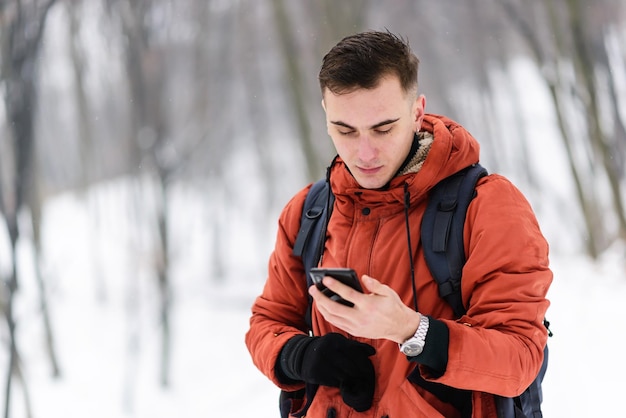 Chico turista mirando su teléfono inteligente en el bosque frío de invierno