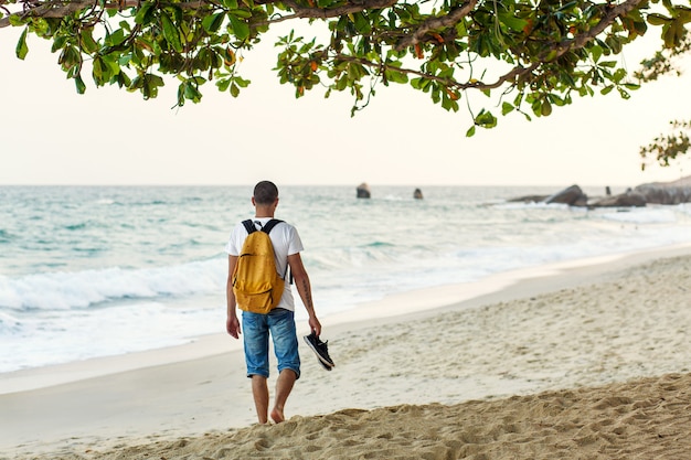 Chico turista con una mochila amarilla camina por la playa de arena junto al océano.