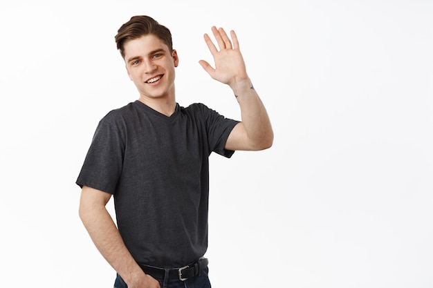 Chico universitario amistoso saluda. Un hombre guapo saludando con la mano levantada para saludarte, hola gesto, sonriendo amablemente, conociendo a alguien, parado sobre fondo blanco