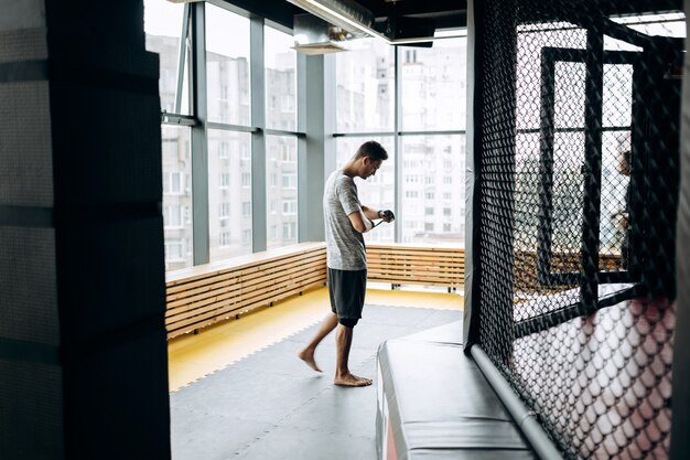 Chico vestido con la camiseta gris envuelve un vendaje en la mano en el gimnasio de boxeo con el telón de fondo de las ventanas panorámicas.