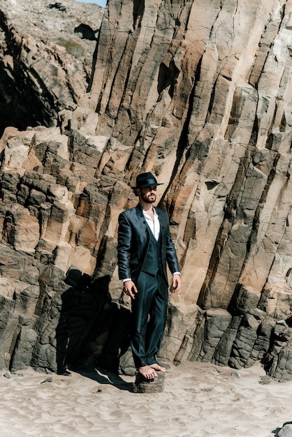 Un chico vestido de novio en una playa con una enorme roca en el fondo