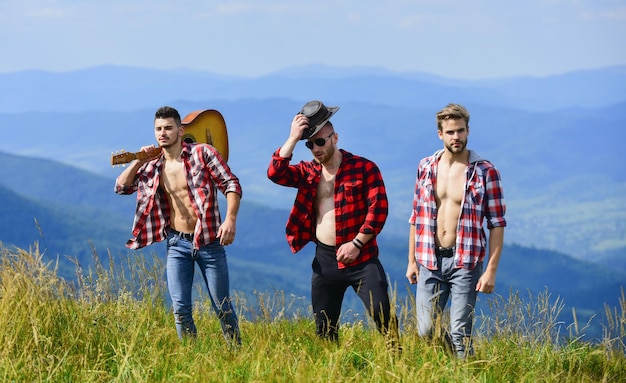 Chicos amables con guitarra haciendo senderismo en un día soleado Disfrutando juntos de la libertad Grupo de jóvenes con camisas a cuadros caminando juntos en la cima de la montaña Concepto de senderismo para turistas Senderismo con amigos