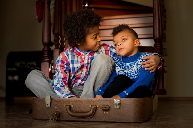 Los chicos amistosos se sientan dentro de la maleta. Dos niños afro de apariencia amigable. Mírame, hermano. Sonríe por la foto, por favor.