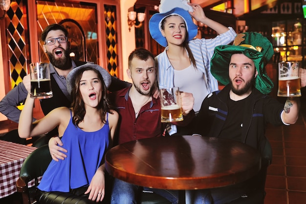 Chicos y chicas jóvenes con vasos de cerveza, viendo fútbol, riendo y sonriendo.