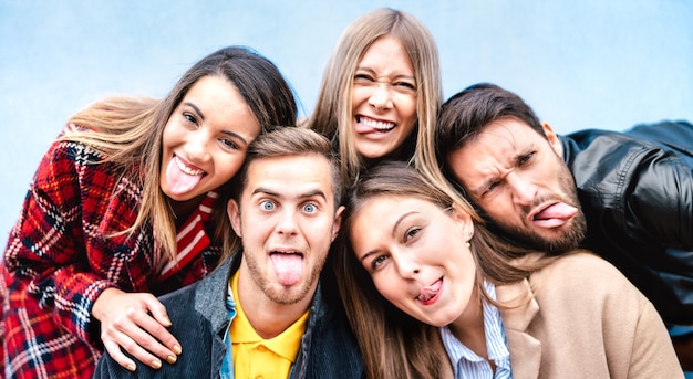 Chicos y chicas milenarios multiculturales tomando selfie sacando la lengua con caras felices