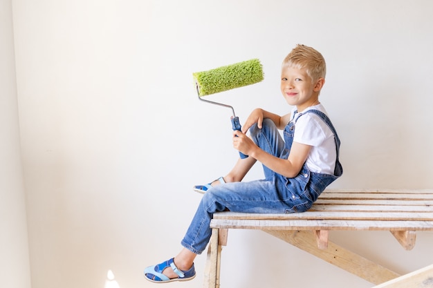 Child boy builder hält bauwerkzeuge in einer wohnung mit weißen wänden, kind malt die wände, platz für text, reparaturkonzept