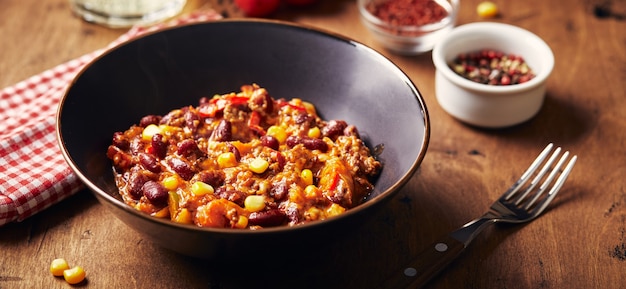 Chili con Carne mit Rinderhackbohnen und Mais in dunkler Schüssel auf Holzhintergrund mexikanisch und texas...