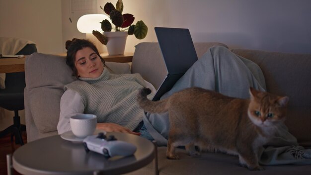 Chilling morena acostada en el sofá viendo la computadora portátil en la sala de noche mujer descansando