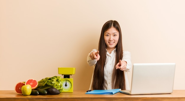 Chinesische Frau der jungen Ernährungswissenschaftlerin, die mit ihrem Laptop fröhliches Lächeln arbeitet, zeigt nach vorne.
