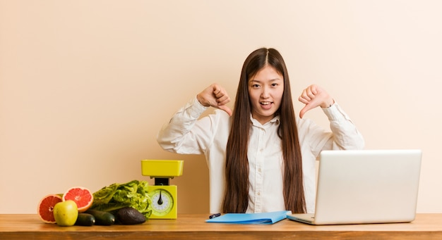 Chinesische Frau des jungen Ernährungswissenschaftlers, die mit ihrem Laptop unten zeigt Daumen und Abneigung ausdrückt.