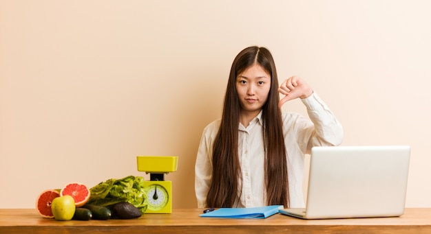 Chinesische Frau des jungen Ernährungswissenschaftlers, die mit ihrem Laptop unten zeigt eine Abneigungsgeste, Daumen arbeitet. Uneinigkeit Konzept.