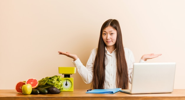 Chinesische Frau des jungen Ernährungswissenschaftlers, die mit ihrem Laptop zweifelt und zuckt mit den Schultern, wenn Geste in Frage gestellt wird.