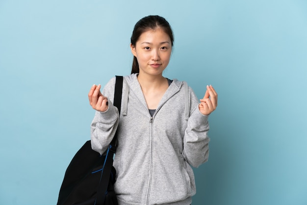 Chinesische Frau des jungen Sports mit Sporttasche über isoliertem Blau, das Geldgeste macht