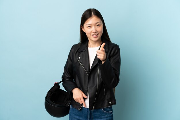 Chinesische Frau, die einen Motorradhelm über blauer Wand hält, zeigt Finger auf Sie