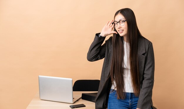 Chinesische Geschäftsfrau an ihrem Arbeitsplatz, der etwas hört, indem man Hand auf das Ohr legt