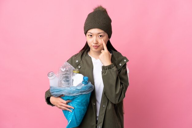 Chinesisches Mädchen, das eine Tasche voller Plastikflaschen hält, um auf lokalisiertem Rosa zu recyceln, das etwas zeigt