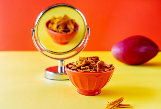 Chips de mango en un tazón con espejo y mango crudo sobre fondo amarillo y naranja