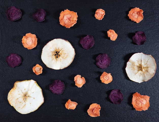 Chips de zanahoria, remolacha y manzana secas sobre un fondo de piedra negra.