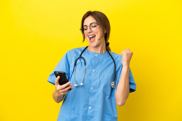 Chirurg Arzt Frau auf gelbem Hintergrund mit Telefon in Siegesposition isoliert