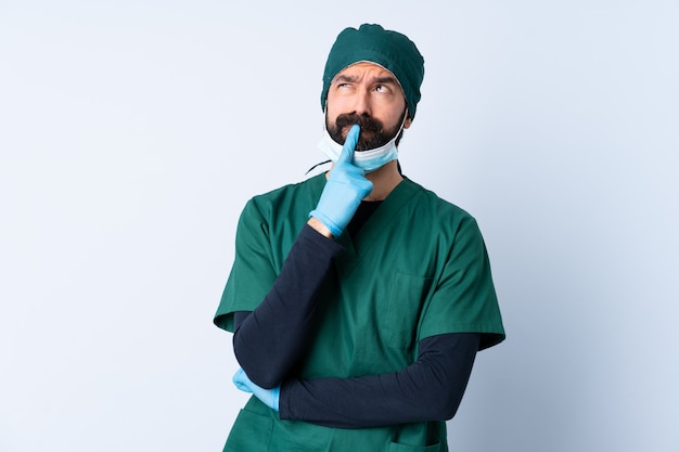 Chirurg Mann in der grünen Uniform über isolierter Wand, die Zweifel beim Aufblicken hat