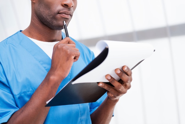 Chirurg mit Zwischenablage. Nahaufnahme eines nachdenklichen jungen afrikanischen Arztes in blauer Uniform, der die Zwischenablage hält und sein Kinn mit einem Stift berührt
